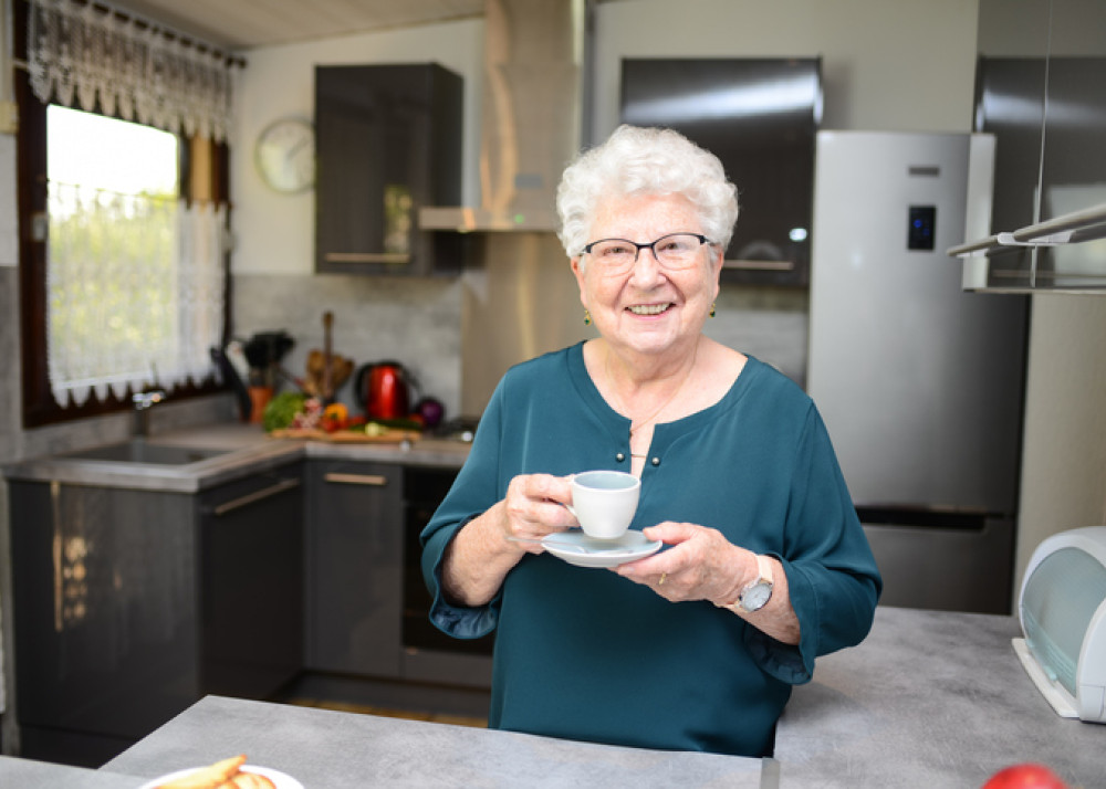 An elderly woman in a teal blouse smiling while holding a cup of tea in her modern kitchen, representing independent living supported by Level 2 Home Care Packages.