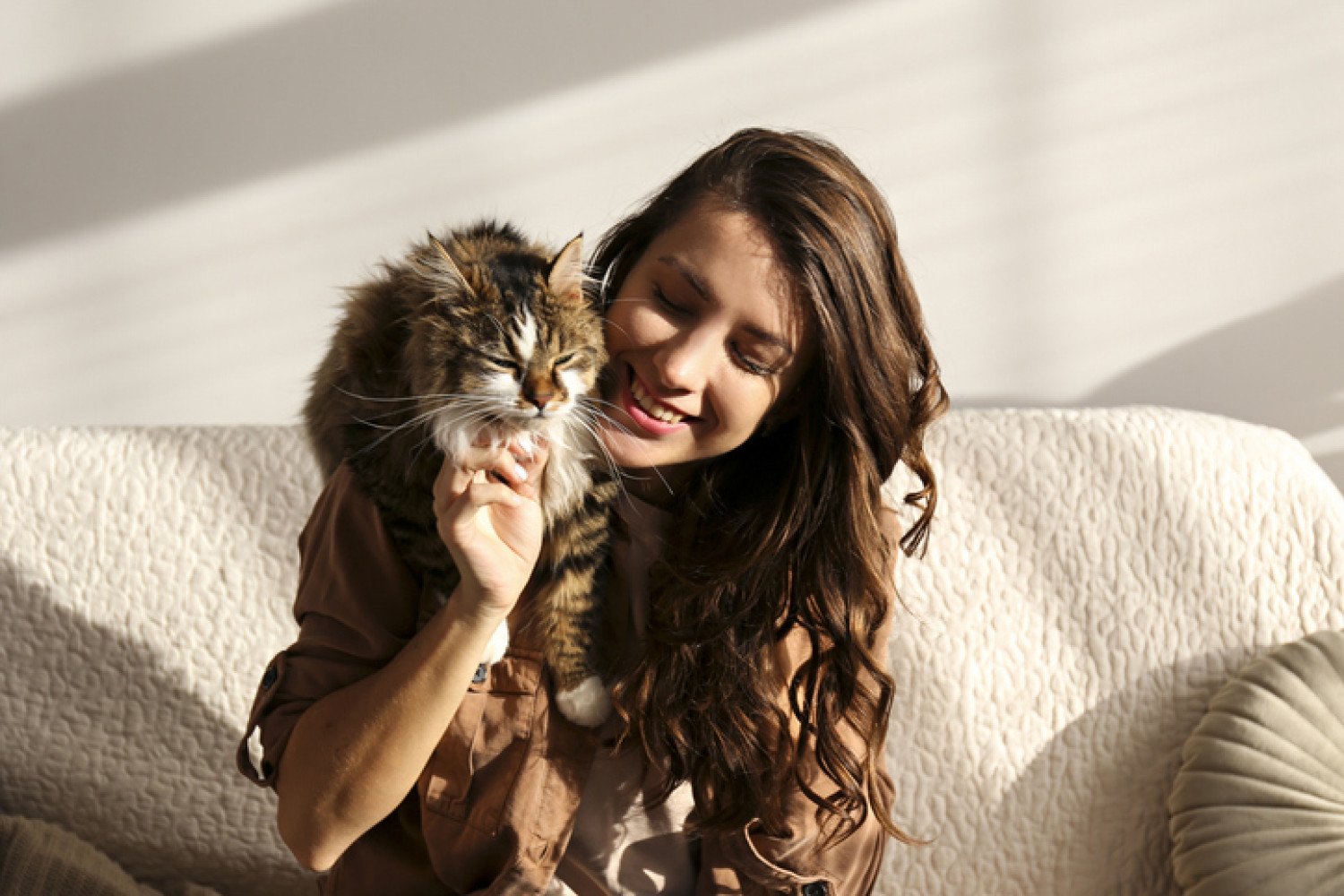 Woman sitting on a sofa petting a cat, in a brightly lit living room.
