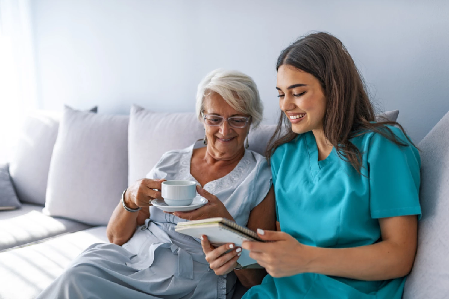 A senior woman in grey pajamas and a blue care worker reviewing writing together on a sofa, depicting the personal and social support offered through Level 2 Home Care Packages.