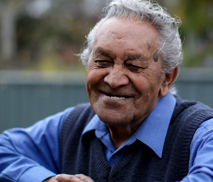 Older indigenous man smiling while leaning on a fence in his backyard.