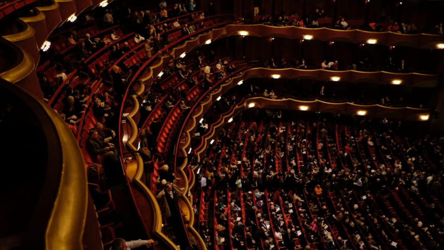 The image shows the inside of the Met Opera in New York City. It looks very extravagant, there are deep red hues and gold colours. There are rows and rows of seats, arranged in an arc shape.