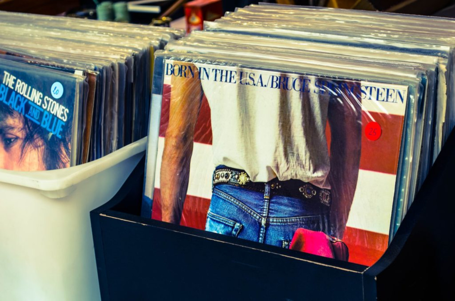 The image depicts two boxes of records. The front record is Born in the USA by Bruce Springsteen. The record cover shows a man with a white T-shirt, a belt and blue jeans with his back to the audience. You can only see his torso, lower back and arms. The background is the red and white stripes of the American flag.