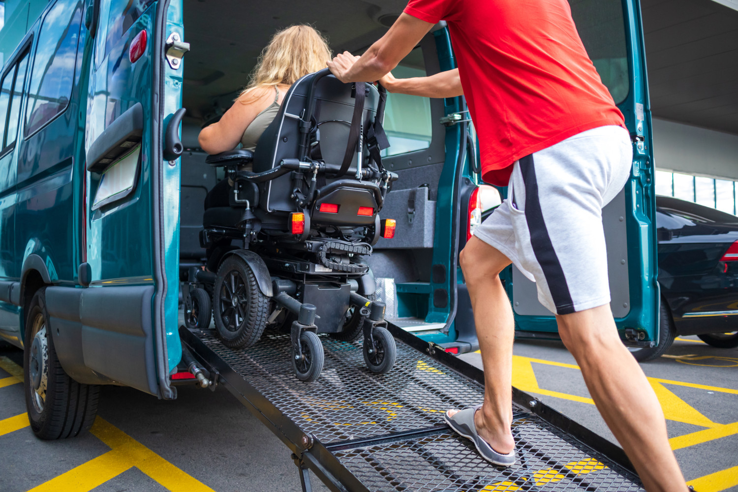 An man is assisting a person in a motorised wheelchair, who is boarding a blue accessible transport vehicle via a deployed ramp.