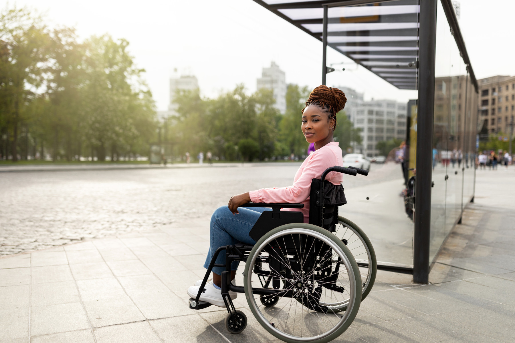 A woman with a confident smile, sits in a wheelchair at a city bus stop waiting for her bus. Representing the accessibility of disability transport services available in Australia.