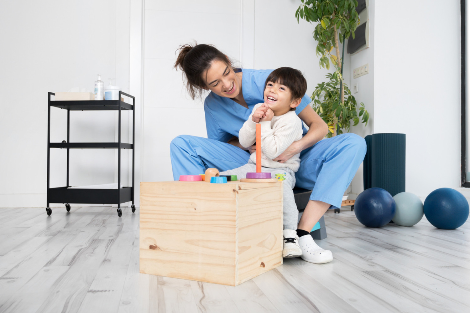 A healthcare professional in blue scrubs affectionately interacting with a young boy while they play with building blocks, symbolizing cerebral palsy support in a therapeutic setting