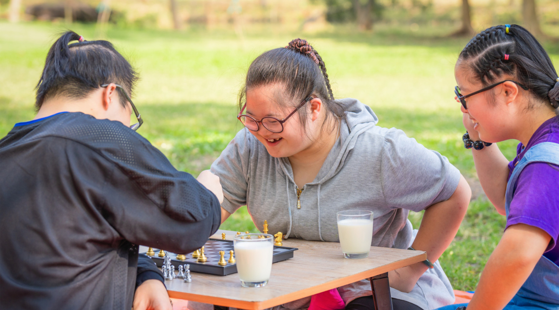 Three individuals enjoying a game of chess outdoors, with a focus on a smiling woman with Down syndrome, highlighting the significance of Mental Health and Disability.