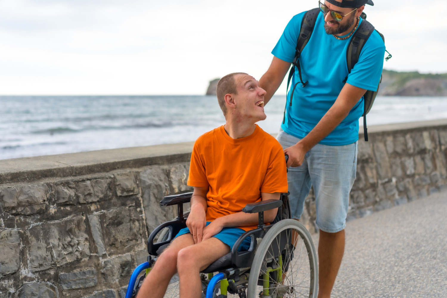 A young man with cerebral palsy in an orange shirt joyfully interacts with a support care worker while seated in his wheelchair against the backdrop of a serene beach.