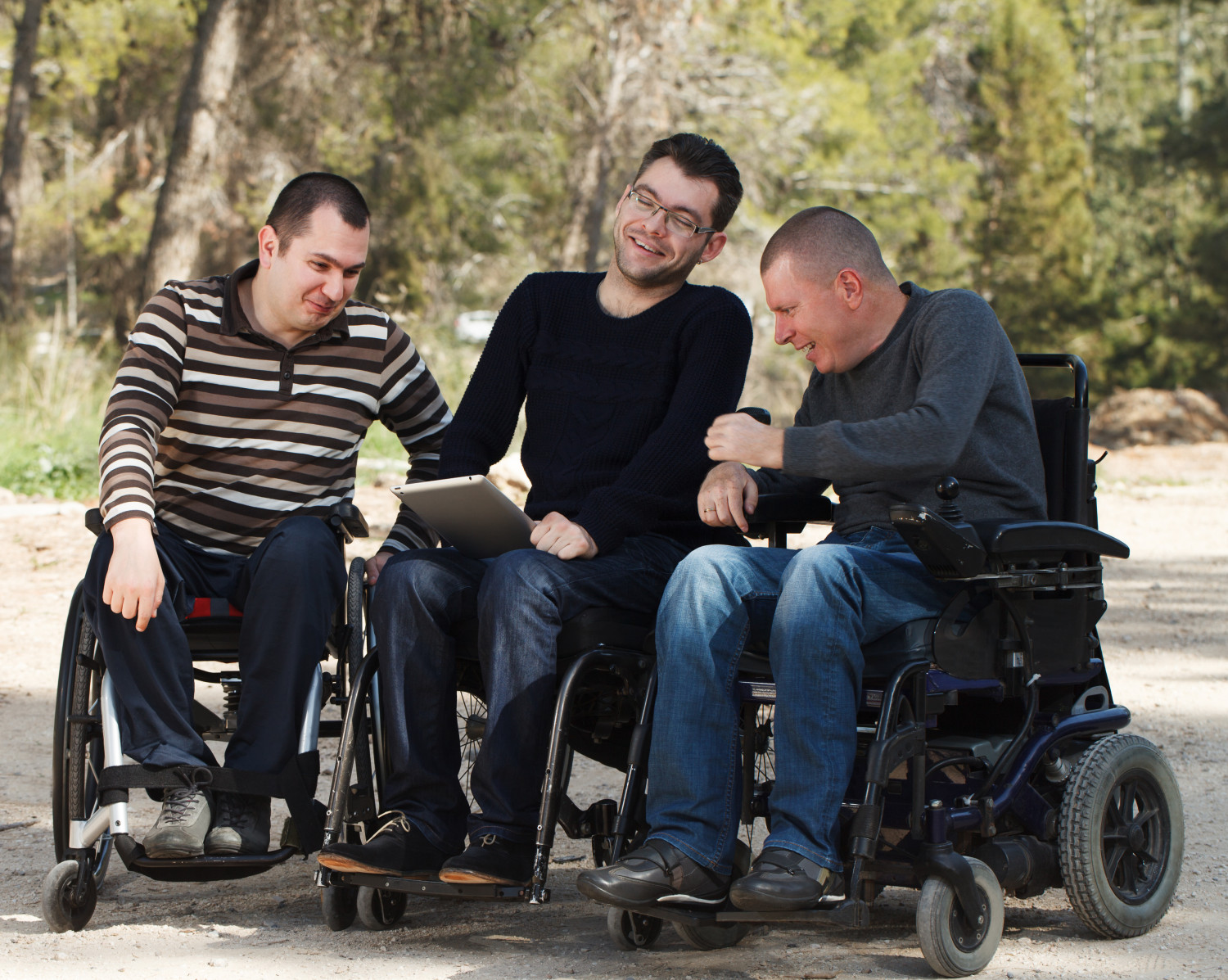 Three men, using wheelchairs, sharing a laugh while looking at a tablet outdoors, showcasing camaraderie in supported independent living