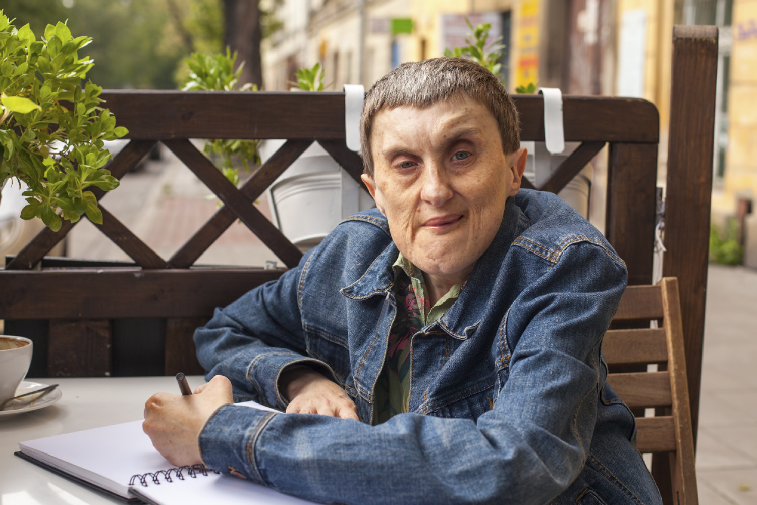 A person with cerebral palsy in a denim jacket sits at an outdoor cafe table, writing in a notebook, exemplifying independence from cerebral palsy support.