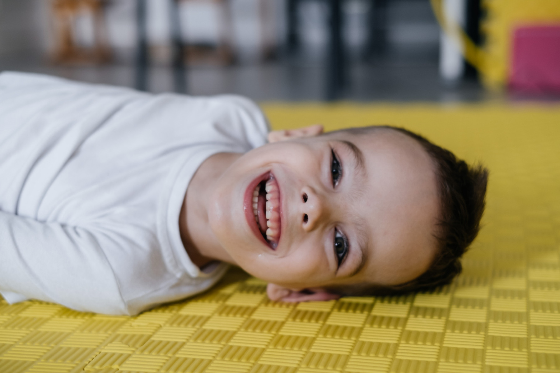 A radiant young boy with cerebral palsy support lays on a yellow mat, gleaming with a cheerful smile.