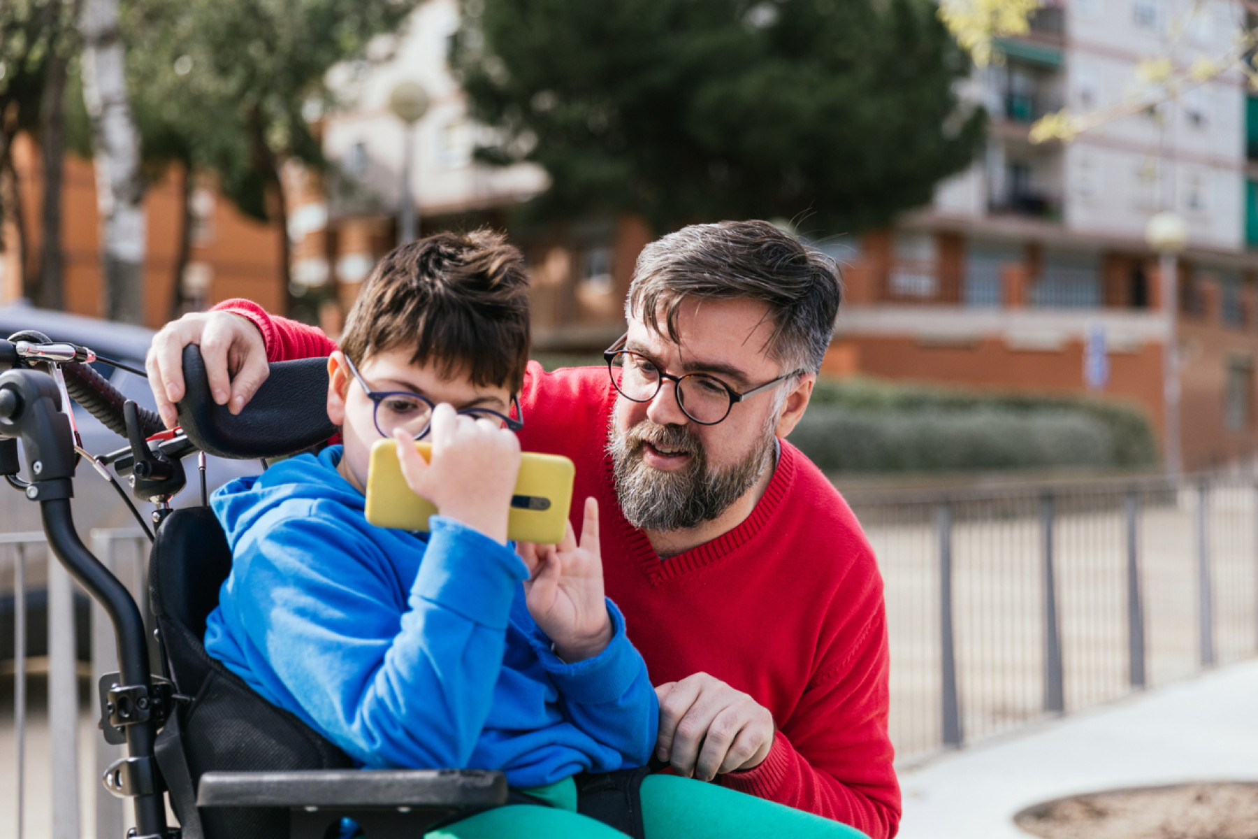 A young boy in glasses seated in a wheelchair using iPhone's Assistive Access, with his father in a red sweater beside him in an outdoor setting.