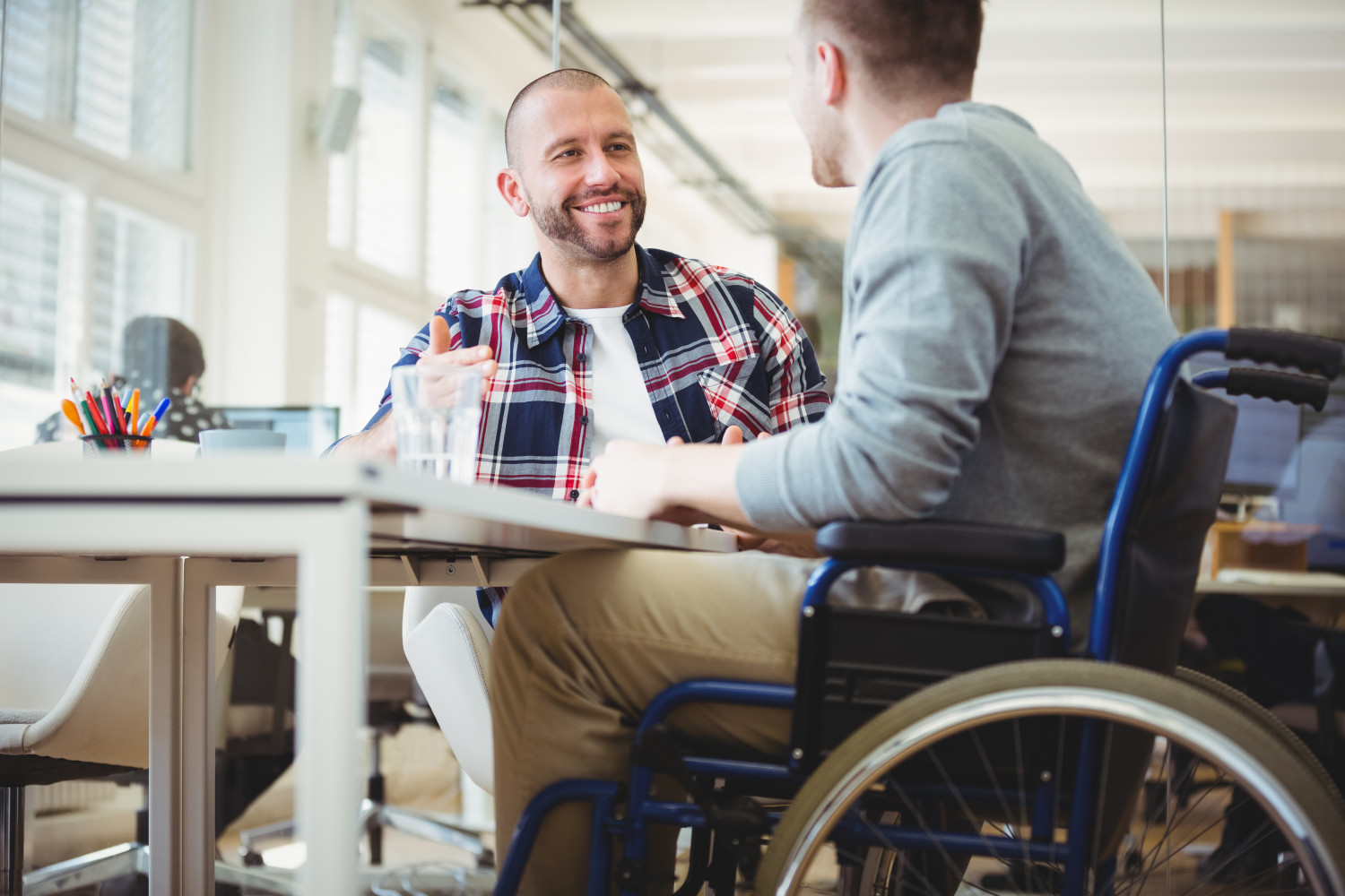 A man in a wheelchair sitting at a desk, smiling at another man - 5 Tips to Know Before Your NDIS Plan Review - Focus Care